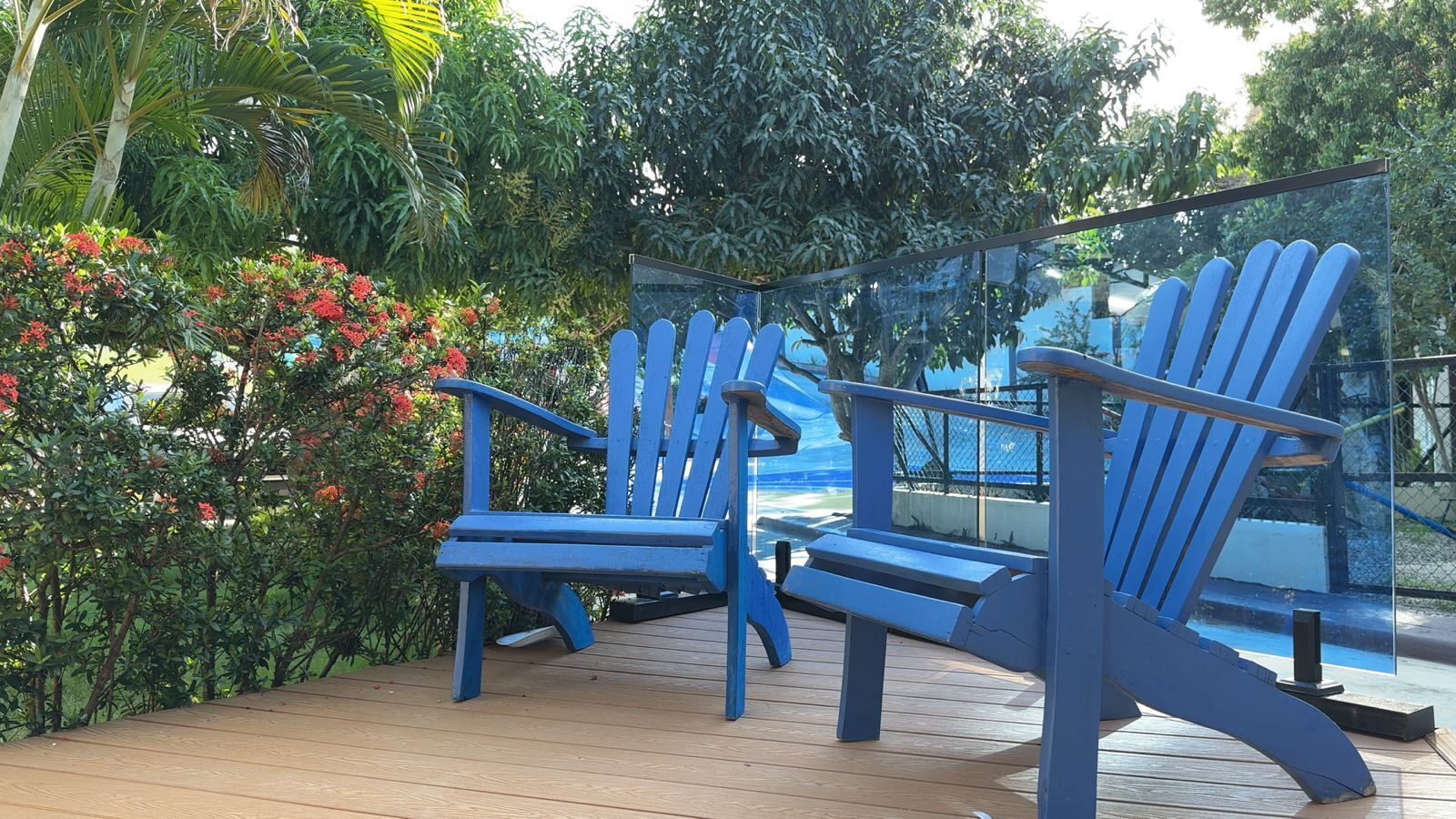 Deux chaises Adirondack bleues sont posées sur une terrasse en bois dotée d’une rambarde en verre, et l’ensemble est entouré d’une végétation luxuriante et de fleurs rouges.
