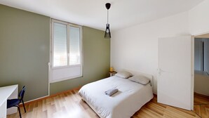 Une chambre à coucher comprenant un lit, une chaise, un bureau, une fenêtre avec des stores et un luminaire suspendu.