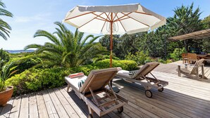 Een houten terras met lounge stoelen en een parasol, omgeven door groen en bomen.