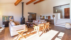 Een ruime woonkamer met een hoge plafond, houten balken, een open haard, een bank, een salontafel en een trap naar een hoger gelegen verdieping.