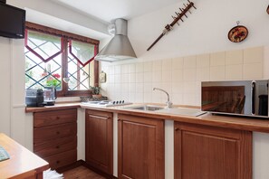 A kitchen with wooden cabinets, a stainless steel sink, a microwave, and a window with a view of greenery. - Petit Logement Cerise "L'écrin d'Ari" – Comfort and Perfect Location (La Cerise)