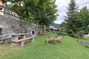 Un muro de piedra con un banco, dos sillas de madera y una pequeña mesa en una zona arbolada con árboles y un edificio al fondo.