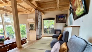 Quarto com esteiras de tatame, sofá com almofadas, TV na parede e vista para o verde lá fora.