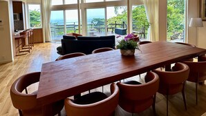 Uma sala de jantar espaçosa com uma longa mesa de madeira e várias cadeiras, grandes janelas com vista para o exterior, e teto com vigas de madeira aparentes e luminárias modernas.