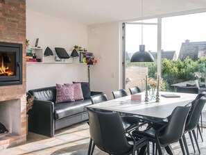 A modern living room with a fireplace, a black leather sofa, a dining table with chairs, and a large window offering a view of the outside. - 4 Star Holiday Home in Romo (Rømø)