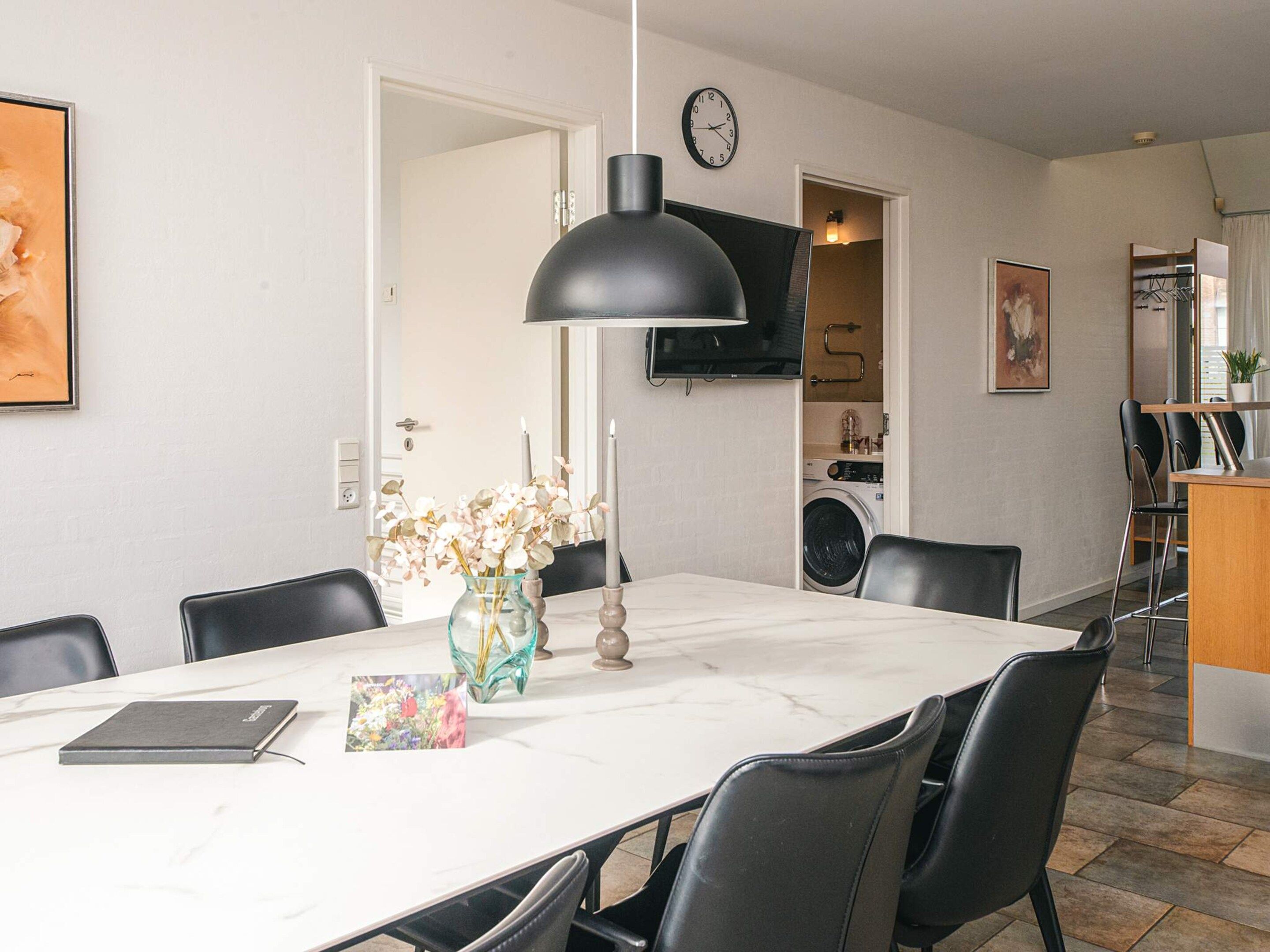 A dining area with a marble table, black chairs, a vase with flowers, a clock on the wall, and a television above the kitchen area.