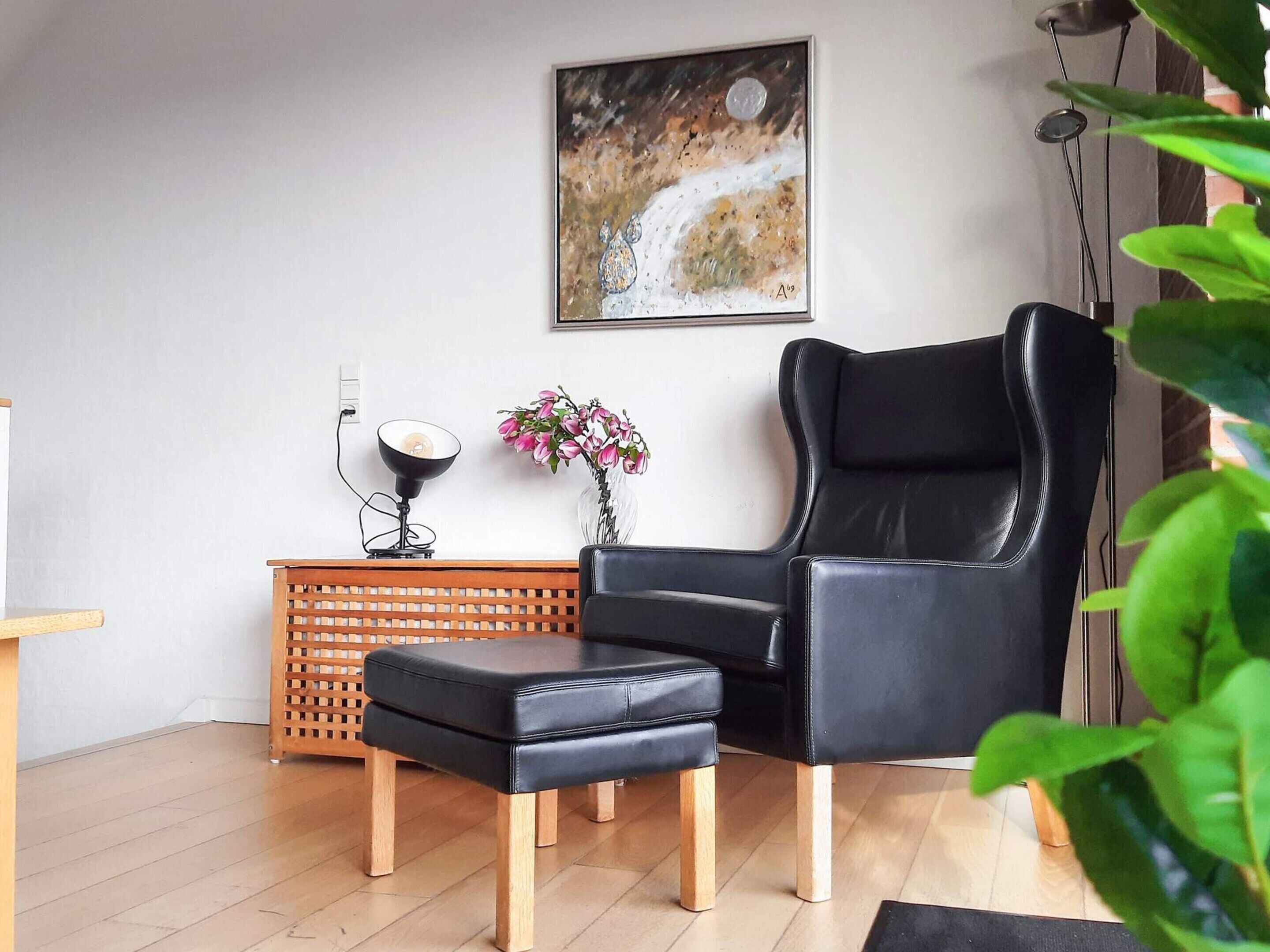 A modern living room with a black leather chair, a wooden side table with a lamp, a vase with flowers, and a painting on the wall.