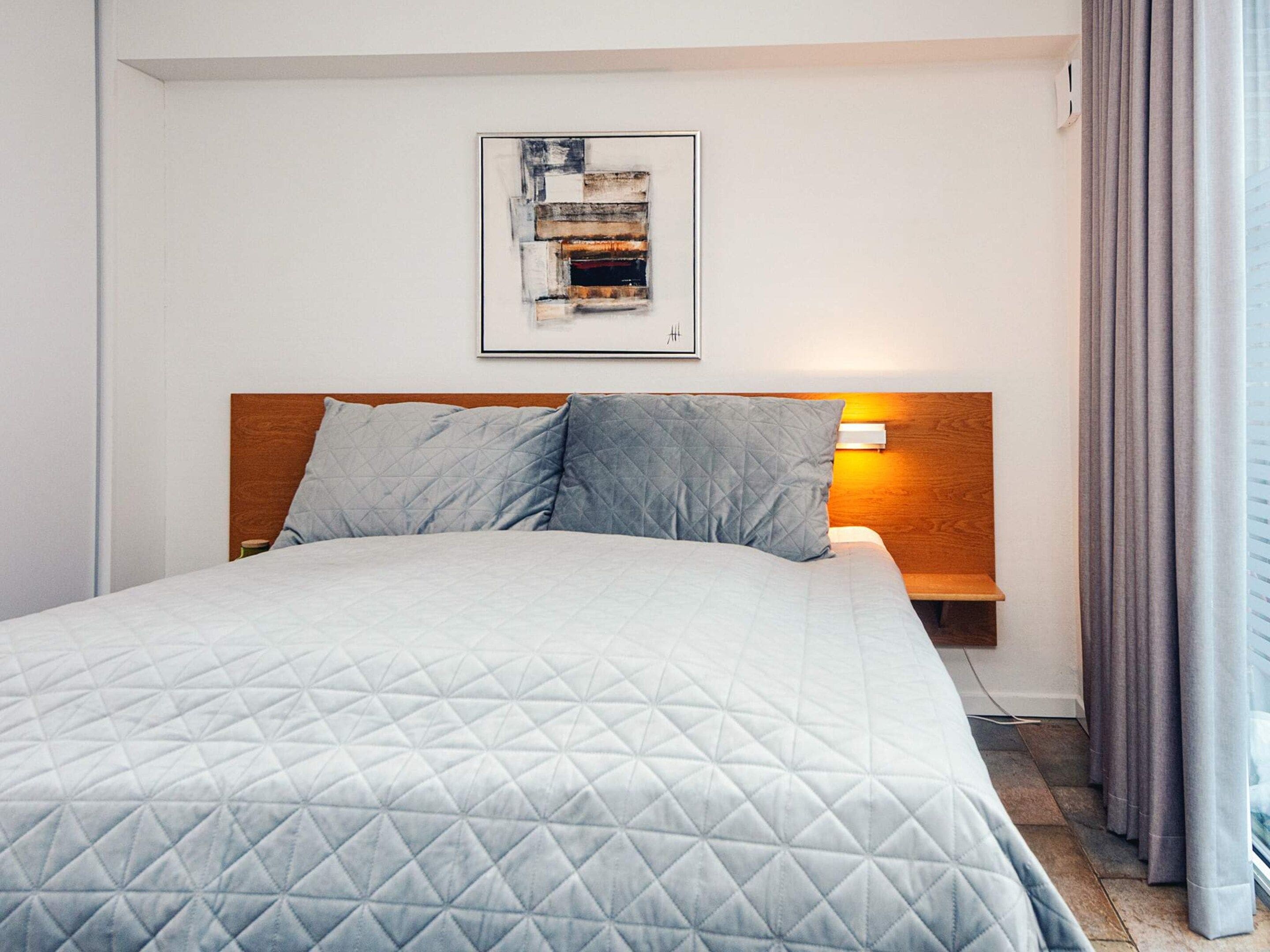 A hotel room with a bed, a headboard, a bedside table with a lamp, and a framed picture on the wall.