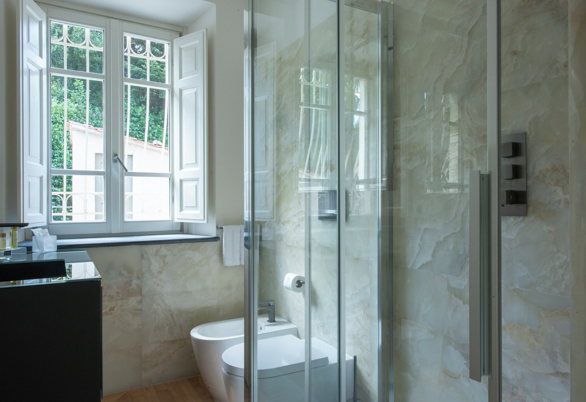 A bathroom with a glass shower enclosure, a toilet, and a window with a view of greenery.