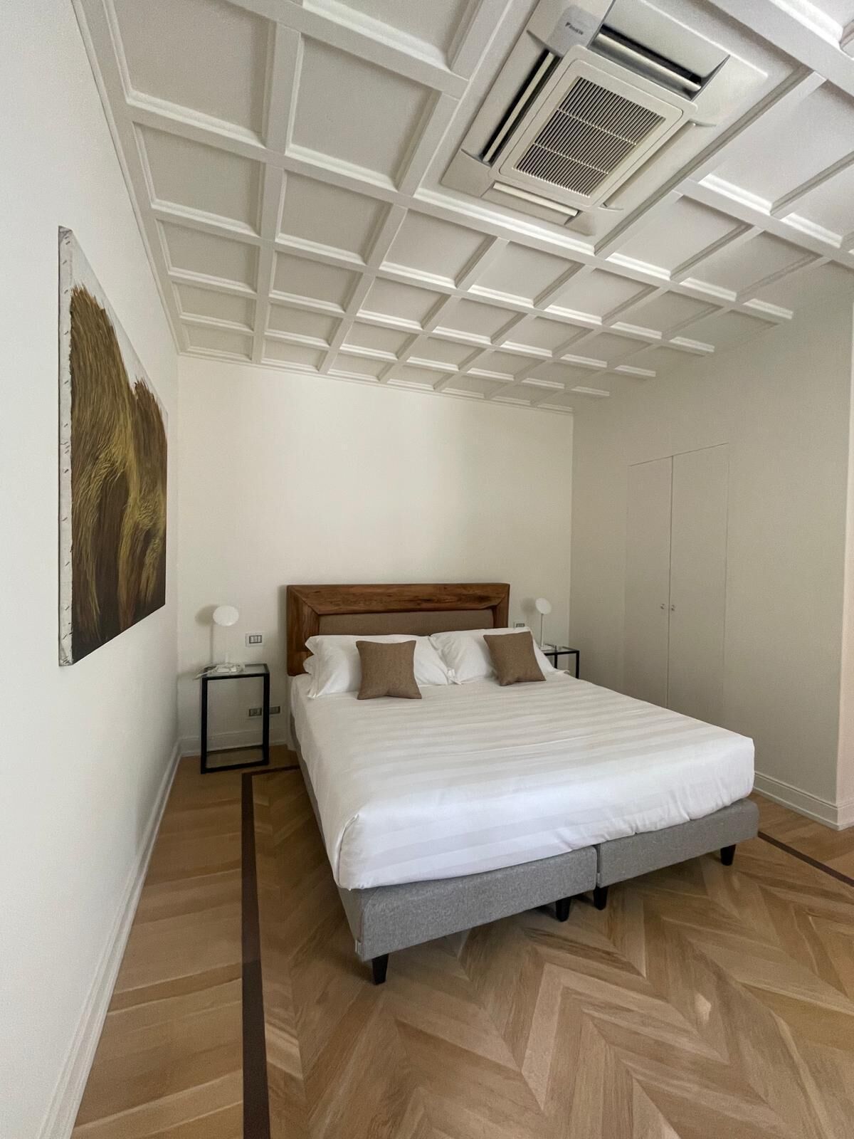 Een slaapkamer met een houten hoofdbord, een groot bed met witte beddengoed, een klein nachtkastje met een lamp en een ventilatie rooster in het plafond.