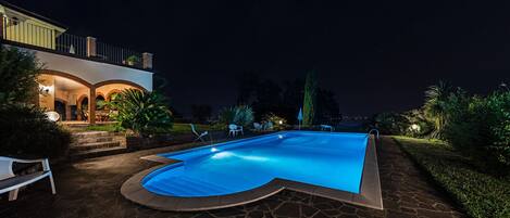 Una piscina con borde curvo, rodeada de baldosas de piedra y un patio con banco y sillas.