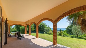 Un patio abrité, agrémenté d’arches en briques, avec une table, des chaises et une vue sur un jardin luxuriant et la mer.