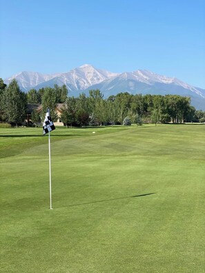 Um campo de golfe com um mastro de bandeira em primeiro plano e uma cordilheira ao fundo.
