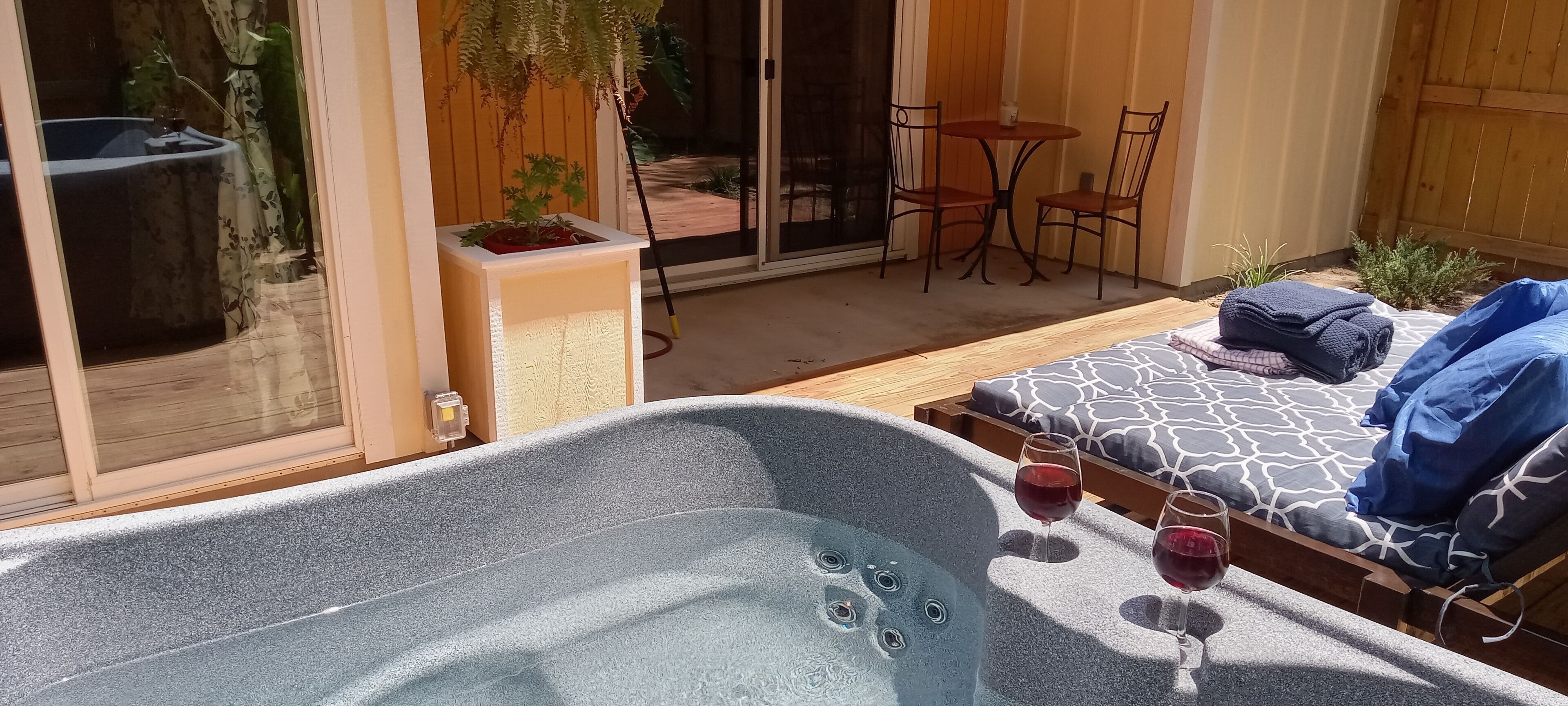 Un bain à remous, deux verres de vin rouge, une terrasse en bois, une table en verre avec des chaises et un coin salon couvert agrémenté d’un coussin à motifs.