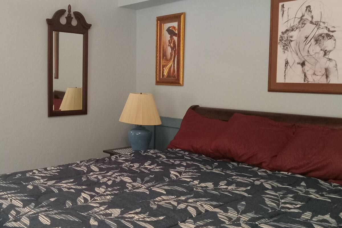 Ein Schlafzimmer mit einem Bett, einem Nachttisch, einer Lampe, einem Spiegel und Kunstwerken an den Wänden.