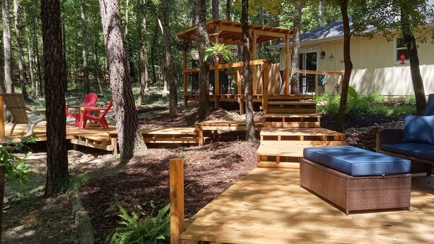 Una terraza de madera con mobiliario exterior, una pérgola y una casa al fondo.