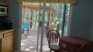 Cocina con microondas, pileta y mostrador de madera. Se ve una mesa con mantel a cuadros rojos y una silla. Fuera, se observa un pérgola de madera y sillas.