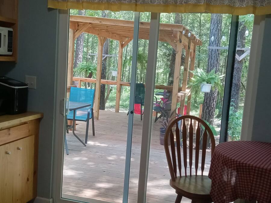 Cuina amb microones, rentapelats i taulell de fusta. Hi ha una taula amb un mantell roig a quadres i una cadira. Se veu una pergola de fusta i cadira a l'exterior.