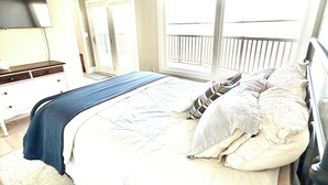 A bedroom with a bed, a nightstand, a dresser, a television, and a window with a view of a balcony.