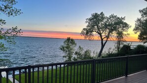 A deck with a view of a sunset over a body of water, surrounded by trees and a grassy area.