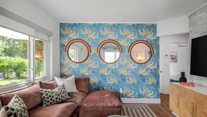 A living room with a brown sectional sofa, a patterned wall, and a TV on a wooden cabinet. - Art Deco Den - Charming 3 bedroom home with pool in Tampa (Tampa)