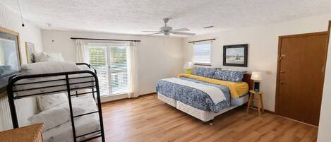 A bedroom with a bunk bed, a single bed, wooden flooring, a ceiling fan, a window with blinds, and a door.
