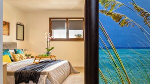 Een slaapkamer met een bed, kussens, een nachtkastje met een lamp, een raam met zonwering en uitzicht op zee.