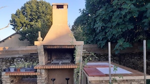 Cucina esterna in pietra con un camino, circondata da piante in vaso e un pontile in legno.