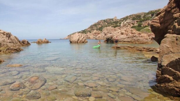 Une zone côtière rocheuse avec une eau peu profonde et claire, une personne sur une planche de paddle verte et des falaises rocheuses.
