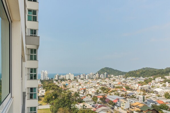 Un immeuble de grande hauteur doté de nombreux balcons, donnant sur une zone urbaine densément peuplée, avec divers bâtiments et une vue lointaine sur la mer.