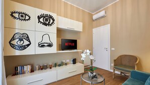 Un salon moderne avec un meuble blanc où sont exposés des objets décoratifs, un téléviseur à écran plat, une petite table avec un vase et un fauteuil en osier.