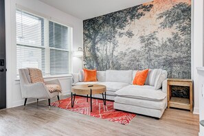 A modern living room with a grey sofa, orange cushions, a round wooden coffee table, and a red patterned rug.