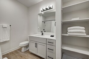 A modern bathroom with a white sink, mirror, and toilet. There are shelves with folded towels and a laundry basket.