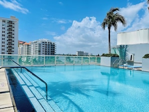 Una piscina azul cristalina con una barrera de cristal, rodeada de edificios modernos y una palmera.