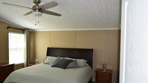 Une chambre à coucher comprenant un lit, une table de chevet, une commode, un ventilateur de plafond et une fenêtre avec des stores.