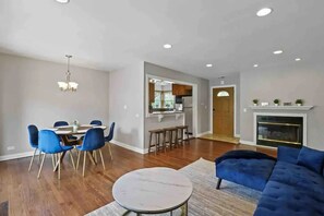 Een ruime woonkamer met een blauwe bank, een eethoek met blauwe stoelen, een keuken met een wit aanrecht en een open haard.