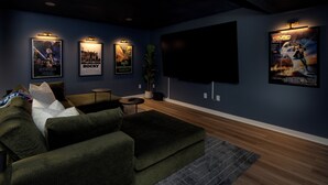 Sala de cinema com uma grande TV de tela plana, um sofá seccional verde, uma mesinha de centro e pôsteres de filmes na parede.