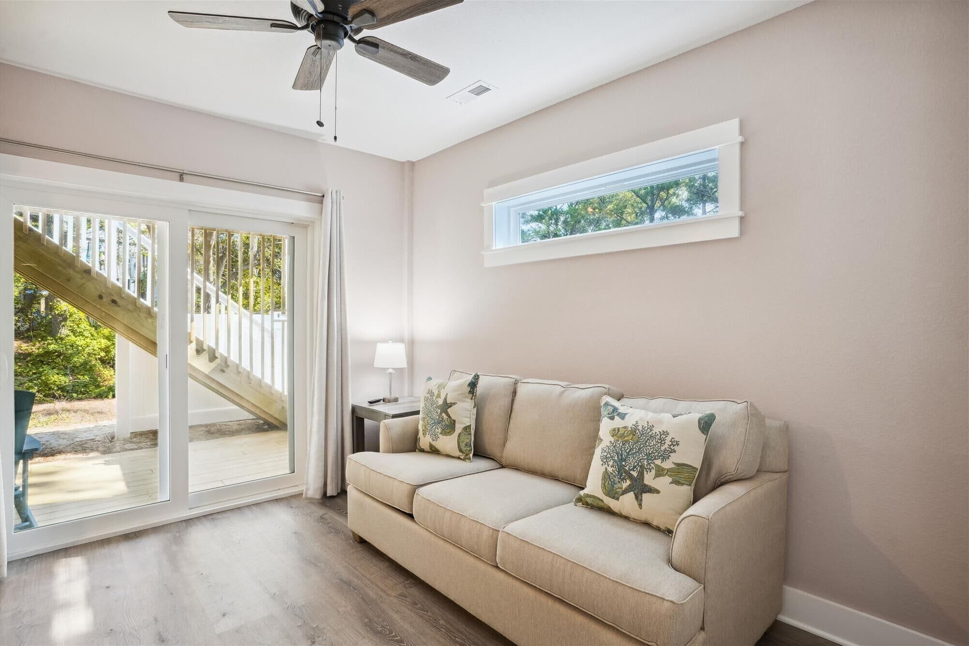 Ein Zimmer mit einem beigefarbenen Sofa, einem Deckenventilator, einem Fenster mit Blick auf Bäume und einer Glastür, die auf eine Holzdiele führt.