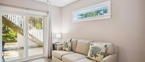 Ein Zimmer mit einem beigefarbenen Sofa, einem Deckenventilator, einem Fenster mit Blick auf Bäume und einer Glastür, die auf eine Holzdiele führt.