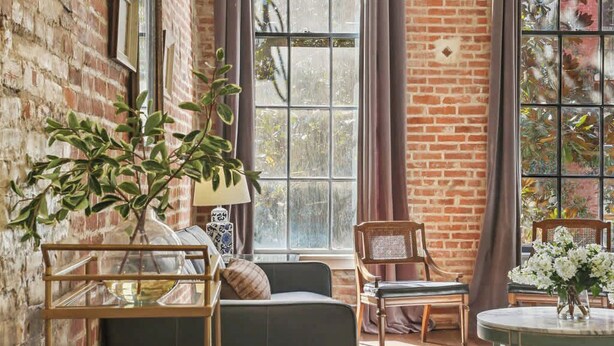 Una habitación con pared de ladrillo, ventanas amplias, un sofá, una silla, una mesa y una planta.