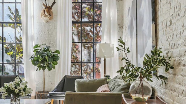 Een kamer met een groot raam, een opgehangen dierenhoofd, een bank, een tafel en een boekenplank.