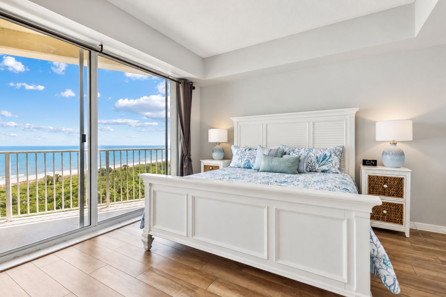 A bedroom with a large bed, two bedside tables, a nightstand, a view of the ocean, and a balcony.
