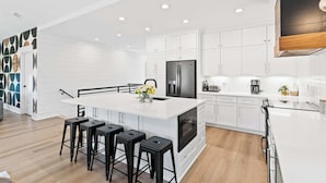 A modern kitchen with white cabinetry, a central island with stools, a built-in oven, and a stainless steel refrigerator. - Modern 6BR Beach Retreat w/ Private Pool (Oak Island)