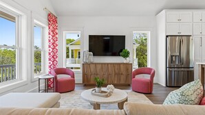 Een moderne woonkamer met een grote televisie, twee roze fauteuils, een houten kast en een keuken met een roestvrijstalen koelkast.