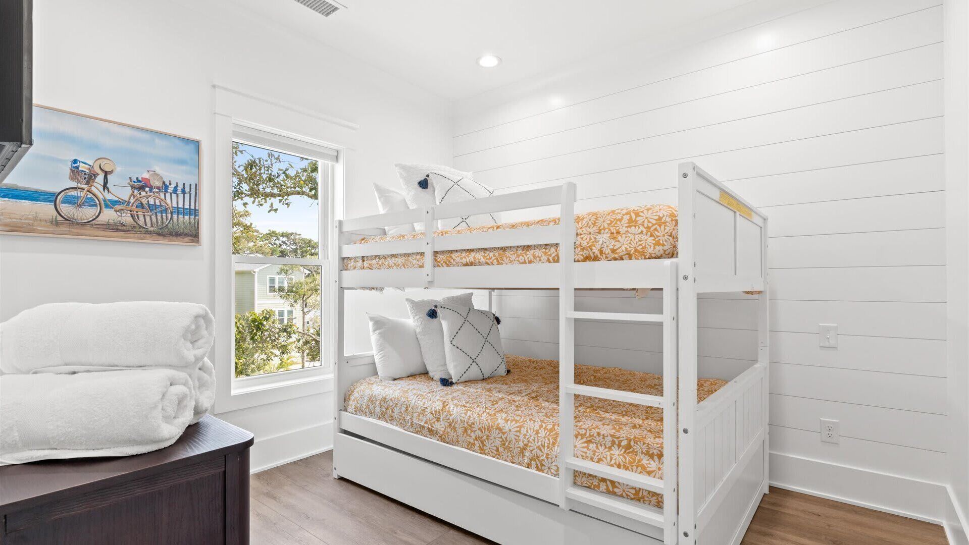 A bunk bed with floral bedding, a window with a view of a house and trees, and a painting of a bicycle on the wall.