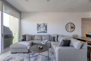 A modern living room with a grey sectional sofa, a small round coffee table, and a large window offering a view of the outdoors. - Modern condo near Medano beach (Cabo San Lucas)