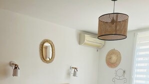 Una camera da letto con una testiera in rattan, un letto grande con molti cuscini, una lampada a sospensione in fibra naturale, un condizionatore d'aria a parete e una decorazione murale raffigurante una donna.