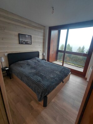 A bedroom with a large bed, wooden flooring, a nightstand, a lamp, and a window with a view of trees. - Le Corbier: a new, cozy apartment right in the heart of the resort! (Villarembert)