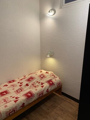 Une petite pièce étroite, avec un lit simple, un mur à texture et un luminaire fixé au mur.