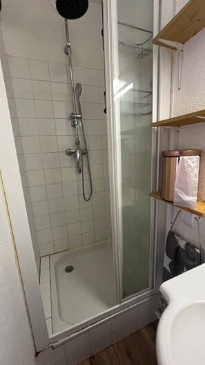 Zona de ducha con mampara de vidrio, pared con azulejos blancos y estante de madera con una toalla.
